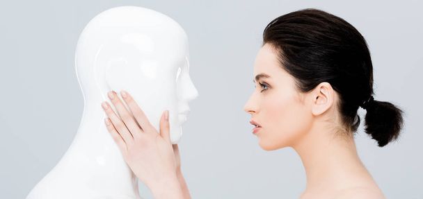 panoramic shot of beautiful young woman touching plastic mannequin isolated on grey - 写真・画像