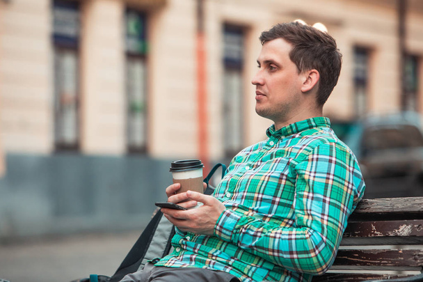Porträt eines jungen gutaussehenden Mannes, der auf einer Bank sitzt und Kaffee trinkt und per Telefon im Internet surft - Foto, Bild