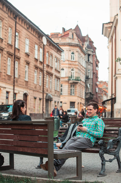 nevetve beszél az ember, nő ül a padon iszik kávét szörfözés az interneten. városi életstílus - Fotó, kép