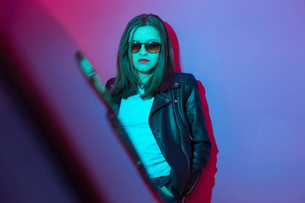 Fashioned girl in leather jacket in dark glasses in pink and blue  neon lights background.  - Foto, afbeelding