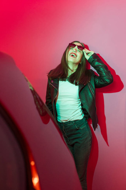 Fashioned girl in leather jacket in dark glasses in pink and blue  neon lights background.  - 写真・画像