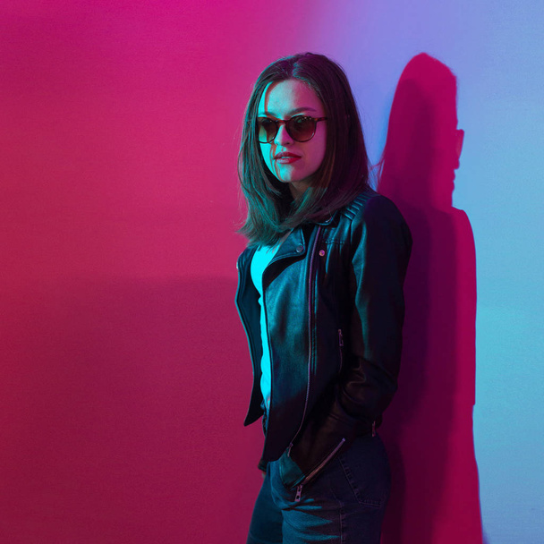 Fashioned girl in leather jacket in dark glasses in pink and blue  neon lights background.  - Foto, afbeelding