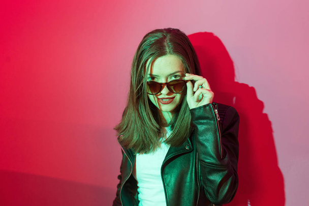Fashioned girl in leather jacket in dark glasses in pink and blue  neon lights background.  - Foto, Imagem