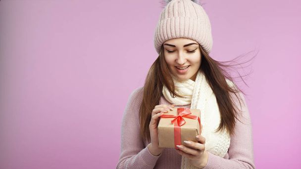 portrait of happy surprised girl opening gift box in decorative craft paper, young woman in knitted hat and scarf on a pink colored background with present, concept of holidays - Photo, image