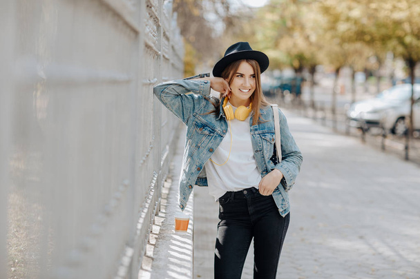 Portrait d'une belle étudiante hipster souriante aux cheveux bruns portant un chapeau, un sac à dos et des écouteurs jaunes dans le parc
. - Photo, image