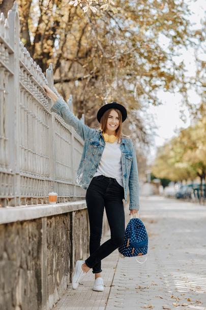 Portrait of a beautiful smiling hipster girl student with brown hair wearing a hat, backpack and yellow headphones in the park. - Photo, image