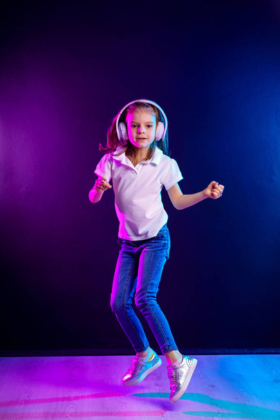 Girl listening to music in headphones on dark colorful background . Dancing girl. Happy small girl dancing to music. Cute child enjoying happy dance music. - Fotó, kép