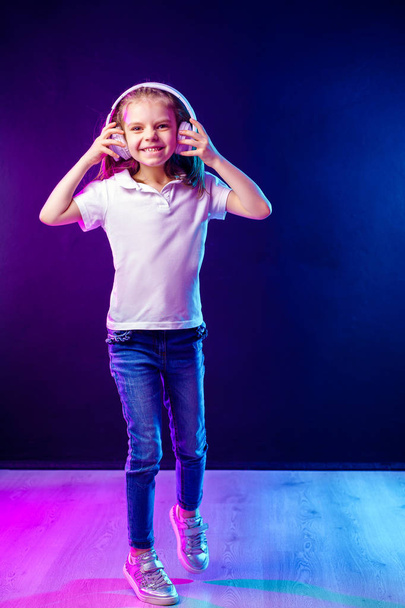 Girl listening to music in headphones on dark colorful background . Dancing girl. Happy small girl dancing to music. Cute child enjoying happy dance music. - Fotó, kép