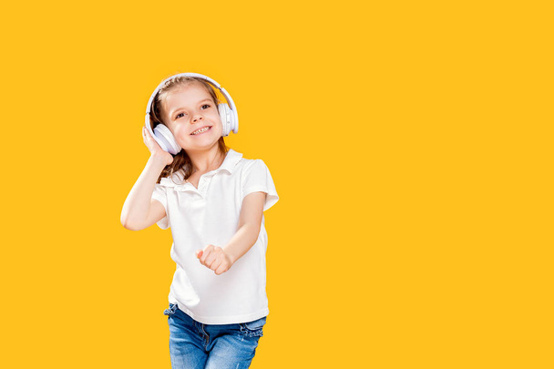 黄色の背景にワイヤレスヘッドフォンで音楽を聴く女の子.踊ってる女の子音楽に踊る幸せな小さな女の子。幸せなダンスミュージックを楽しむかわいい子供. - 写真・画像