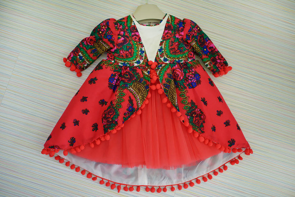 Απομονωμένο αξιολάτρευτο φόρεμα για μωρά κορίτσια φτιαγμένα από κόκκινο βαμβάκι ή κακαμμένο βαμβάκι, με λαχούρι πολύχρωμο αποτύπωμα στο στυλ των σάλια του Pavpόβο Posad, ένα παραδοσιακό ρωσικό μοτίβο μόδας, και με φούστα από κόκκινο τούλι - Φωτογραφία, εικόνα
