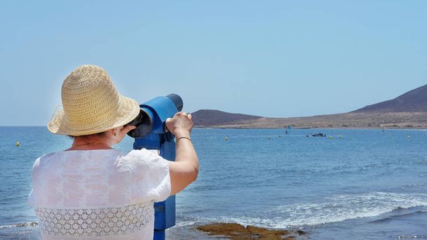 İspanya 'nın Kanarya Adaları, El Madano' da güneşli bir günde, yaz tatili boyunca, şapka takan ve elinde mavi teleskop dürbünü tutan yaşlı kadın yolcuların arka görüntüsü. - Fotoğraf, Görsel