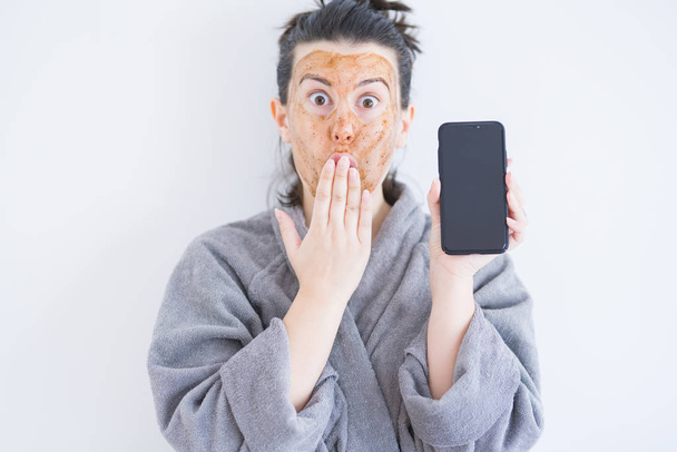 Beautiful woman wearing cosmetic facial mask as skincare treatment showing smartphone screen cover mouth with hand shocked with shame for mistake, expression of fear, scared in silence, secret concept - Photo, Image