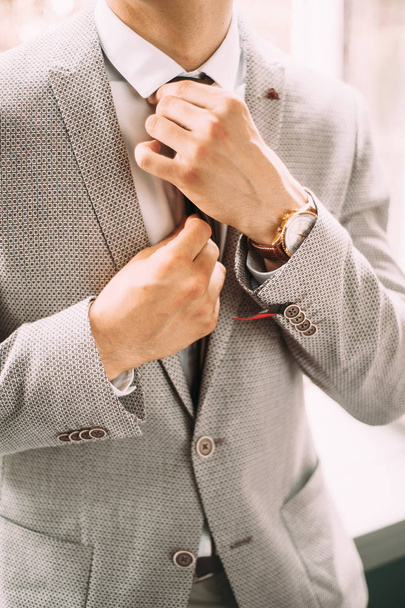 Ανδρική μόδα. Όμορφος άνδρας φορώντας κομψό γκρι κοστούμι και ρολόι χειρός ισιώνει μια γραβάτα - Φωτογραφία, εικόνα