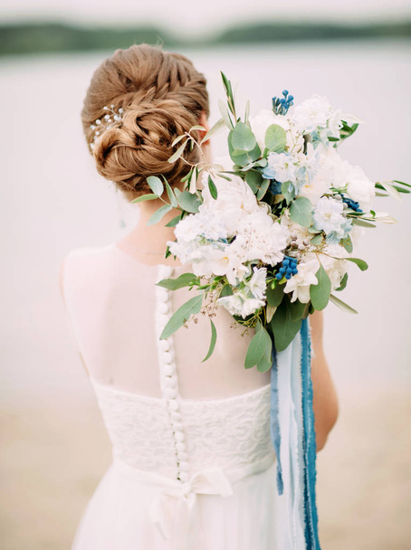 結婚式の日。ウェディングドレスの花嫁は、海のビーチの近くに白と青の花で柔らかいブライダルブーケを保持しています - 写真・画像
