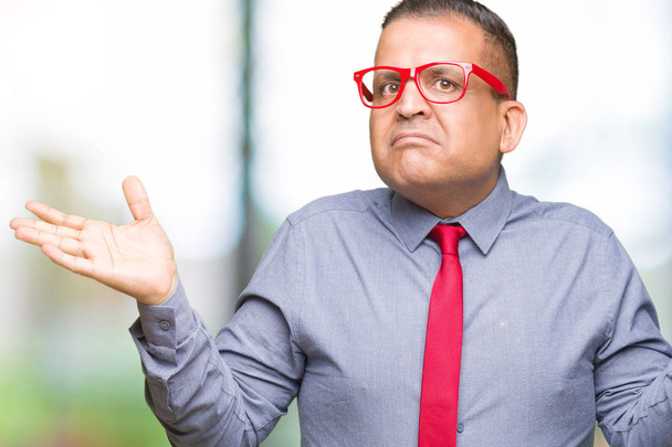 Homem árabe de meia idade vestindo moda óculos vermelhos sobre fundo isolado indolente e confuso expressão com braços e mãos levantadas. Conceito de dúvida
. - Foto, Imagem