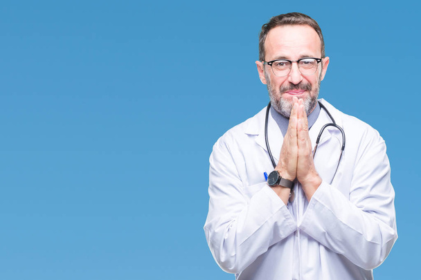Middle age senior hoary doctor man wearing medical uniform isolated background praying with hands together asking for forgiveness smiling confident. - Valokuva, kuva