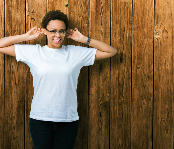 Linda jovem afro-americana vestindo óculos sobre fundo isolado Sorrindo puxando orelhas com os dedos, gesto engraçado. Problema de audição
 - Foto, Imagem