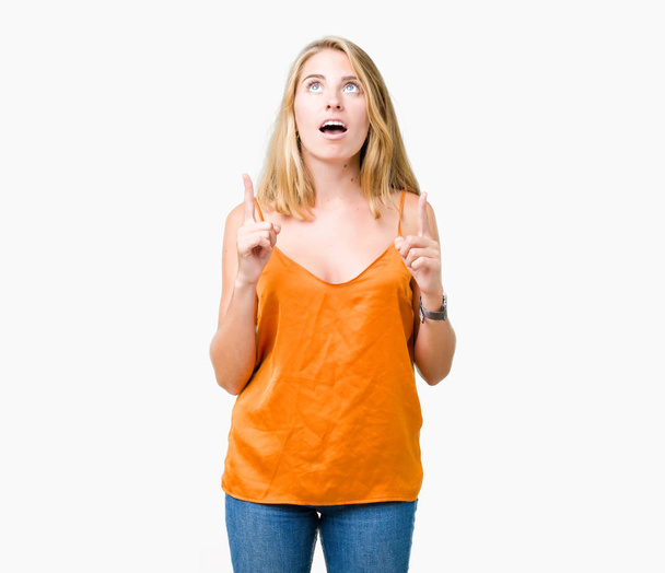 schöne junge Frau, die orangefarbenes Hemd über isoliertem Hintergrund trägt, erstaunt und überrascht nach oben schaut und mit erhobenen Fingern und Armen zeigt. - Foto, Bild