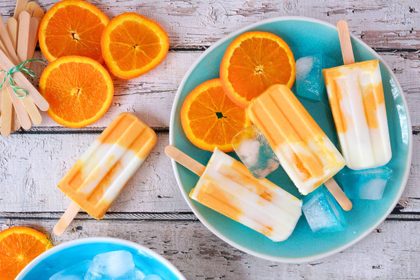 Orange joghurt popsicles egy kék lemez, felülnézet asztali jelenet ellen, fehér fa háttér - Fotó, kép
