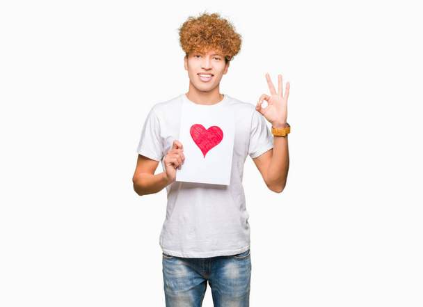 Jovem homem bonito segurando papel com coração vermelho fazendo sinal ok com os dedos, excelente símbolo
 - Foto, Imagem