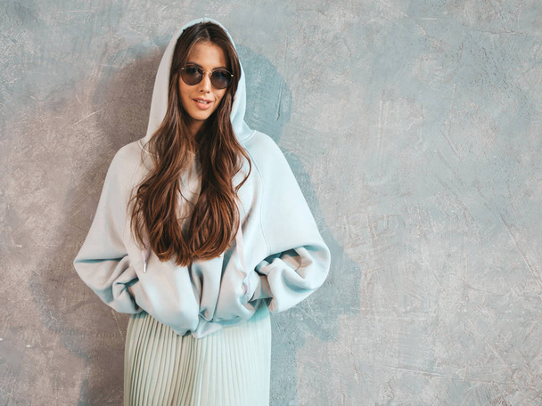 Young beautiful smiling woman looking at camera. Trendy girl in casual summer hoodie and skirt clothes. Funny and positive female posing near gray wall in studio. In sunglasses - Photo, image