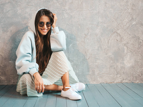 Young beautiful smiling woman looking at camera. Trendy girl in casual summer hoodie and skirt clothes. Funny and positive female posing near gray wall in studio. In sunglasses sitting on the floor - Foto, afbeelding