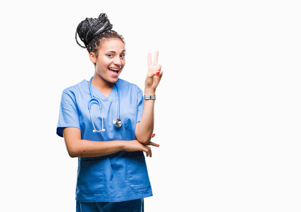 Młody pleciony włosy african american girl profesjonalnej chirurga na białym tle uśmiechnięty z szczęśliwy twarz mrugając aparat robi znak zwycięstwa. Numer dwa. - Zdjęcie, obraz