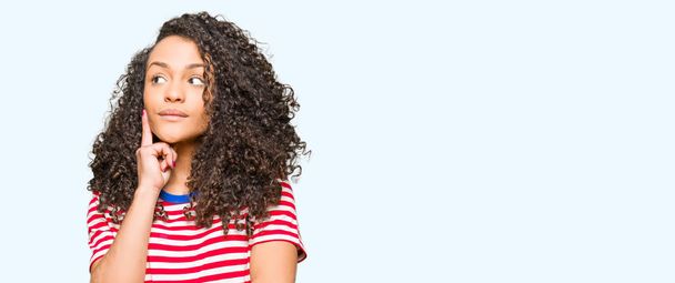 Young beautiful woman with curly hair wearing stripes t-shirt with hand on chin thinking about question, pensive expression. Smiling with thoughtful face. Doubt concept. - Photo, Image