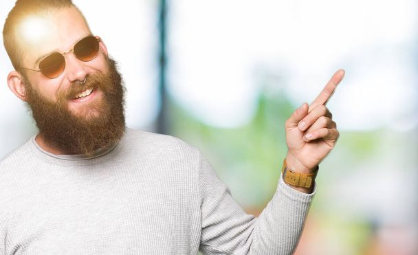 Giovane hipster uomo indossa occhiali da sole con un grande sorriso sul viso, puntando con mano e dito verso il lato guardando la fotocamera
. - Foto, immagini