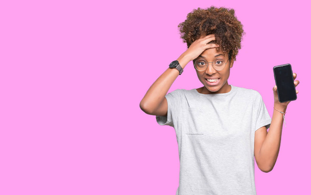 Mujer afroamericana joven mostrando la pantalla del teléfono inteligente sobre un fondo aislado estresado con la mano en la cabeza, conmocionado con la vergüenza y la cara sorpresa, enojado y frustrado. Miedo y malestar por error
. - Foto, Imagen