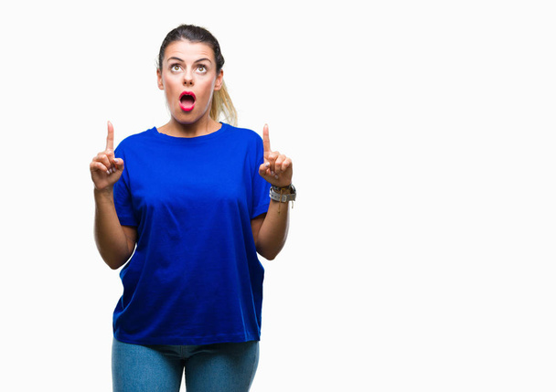 Young beautiful woman wearing casual blue t-shirt over isolated background amazed and surprised looking up and pointing with fingers and raised arms. - Photo, Image