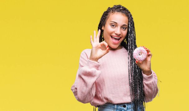 Young african american girl eating pink donut over isolated background doing ok sign with fingers, excellent symbol - Foto, afbeelding