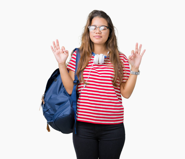 Молодая красивая брюнетка студентка в наушниках и рюкзаке на изолированном фоне расслабляется и улыбается с закрытыми глазами, делая жест медитации пальцами. Концепция йоги
. - Фото, изображение