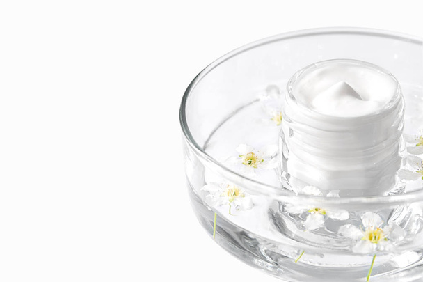A glass jar with cream is in a glass transparent container with water and flowers - Photo, Image