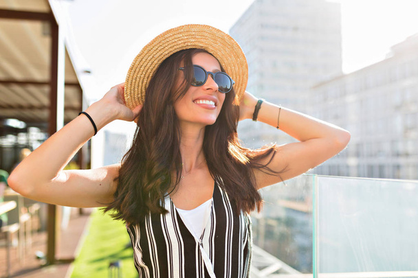 Крупним планом портрет приємної білої жінки в окулярах, що торкається її капелюха на міському фоні на сонячному світлі
. - Фото, зображення