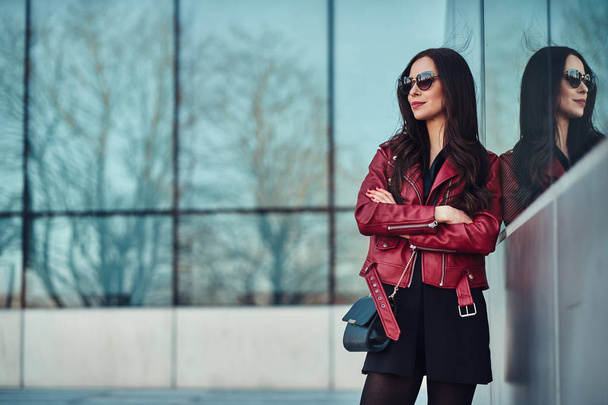 Attraente donna sorridente in giacca rossa e occhiali da sole è in posa vicino edificio di vetro
 - Foto, immagini