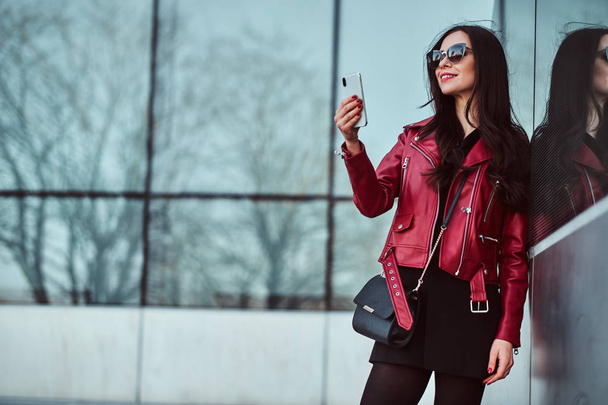 Kırmızı ceketli ve güneş gözlüklü mutlu gülümseyen kadın cep telefonuyla selfie çekerken dışarıda yürümenin tadını çıkarıyor - Fotoğraf, Görsel