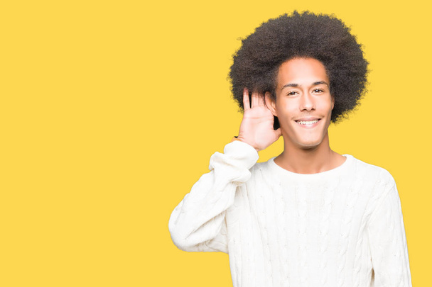 Young african american man with afro hair wearing winter sweater smiling with hand over ear listening an hearing to rumor or gossip. Deafness concept. - Foto, Bild