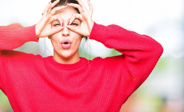 Young beautiful woman wearing red sweater and bun doing ok gesture like binoculars sticking tongue out, eyes looking through fingers. Crazy expression. - 写真・画像