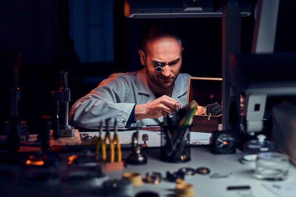集中した男性は、古い時計を修理するモノクルや他のツールで彼のワークショップで働いています - 写真・画像