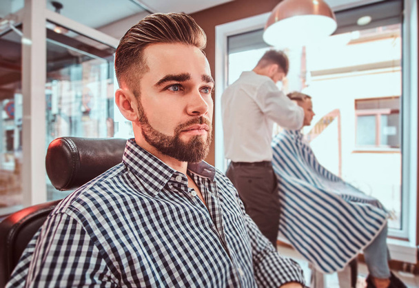 Groomed handsome man is waiting for his turn to get a haircut at busy barbershop - Foto, imagen