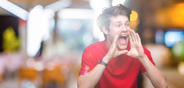 Молодой красивый мужчина в красной футболке на изолированном фоне кричит громко, с руками над ртом
 - Фото, изображение
