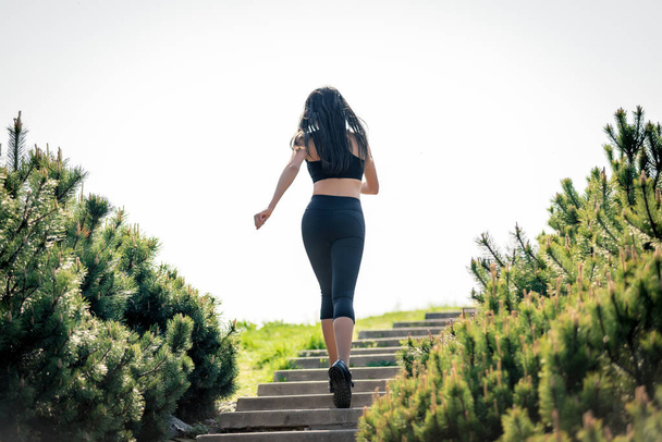 Λεπτό όμορφο κορίτσι με σκούρα μαλλιά τρέχει πάνω από τις σκάλες, ένα σύμβολο του υγιεινού τρόπου ζωής, ένα ισχυρό ηγέτη και υγιή ανταγωνισμό - Φωτογραφία, εικόνα