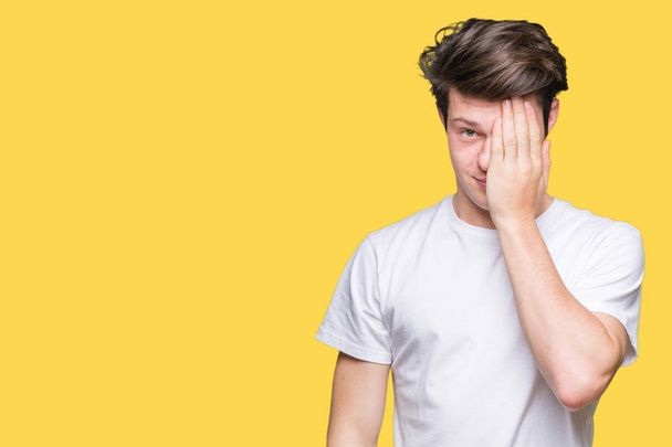 Young handsome man wearing casual white t-shirt over isolated background covering one eye with hand with confident smile on face and surprise emotion. - Photo, Image