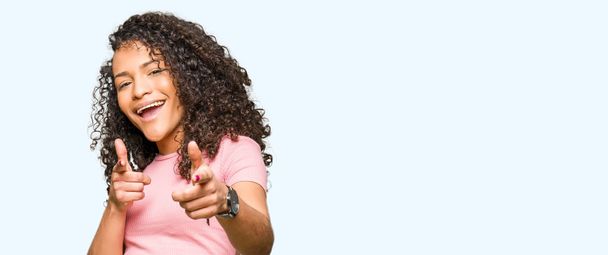 Nuori kaunis nainen kihara tukka yllään vaaleanpunainen t-paita osoittaa sormella kameraan onnellinen ja hauska kasvot. Hyvä energia ja vibes
. - Valokuva, kuva