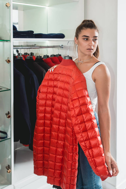 Young woman in a blu jeance white t-shirt trying on red down coat in a luxury boutique - Fotoğraf, Görsel