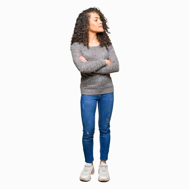 Young beautiful woman with curly hair wearing grey sweater smiling looking to the side with arms crossed convinced and confident - Foto, Imagen
