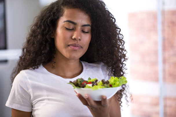 χαρούμενος νεαρός Αφρο αμερικανική γυναίκα τρώει σαλάτα λαχανικών στην κουζίνα στο σπίτι. - Φωτογραφία, εικόνα