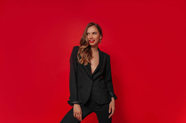 Uśmiechnięty atrakcyjny stylowy model ubrany w czarny kombinezon z faliste włosy i czerwone usta stwarzające w Studio - Zdjęcie, obraz