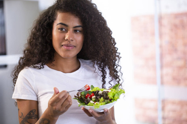 家庭の台所で野菜サラダを食べて陽気な若いアフロ ・ アメリカ人女性. - 写真・画像